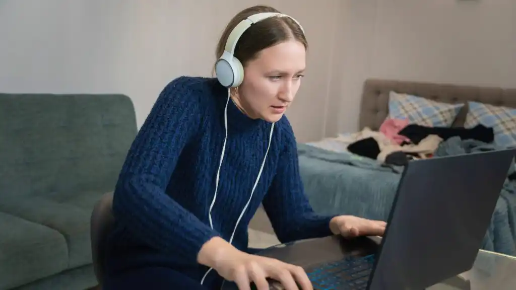 Mujer estudiando a distancia con audífonos frente a una laptop, en una habitación desordenada con ropa y objetos fuera de lugar, reflejando los desafíos de la educación a distancia en la actualidad y la dificultad para concentrarse en entornos no estructurados.