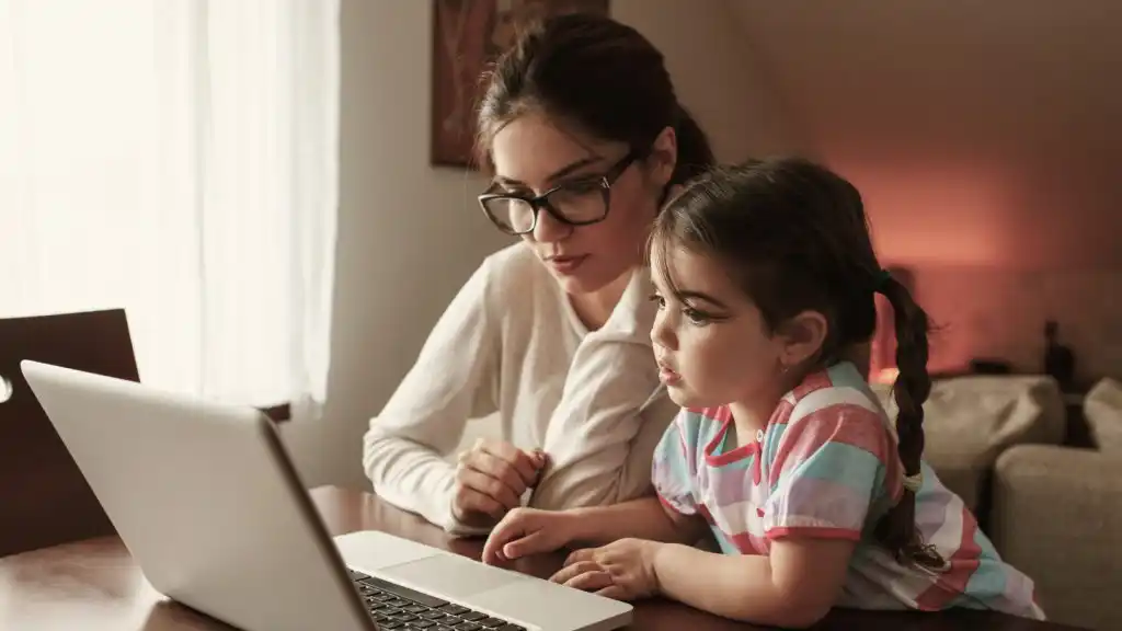Madre joven estudia en casa junto a su hija pequeña frente a una laptop, reflejando el desafío de conciliar la maternidad y trabajo en entornos de aprendizaje digital.